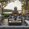 A covered porch overlooks a calm bay at twilight, featuring a graphite patio fire pit surrounded by cushioned armchairs. The steady flames of the fire feature reflect on the stone floor, creating a cozy backyard fire pit atmosphere. This propane fire pit setup provides warmth and a focal point for evening relaxation by the water.