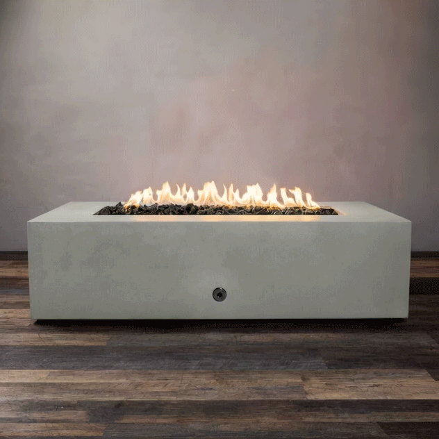 A studio presentation highlights a substantial rectangular fire pit in a natural gray finish, resting on a dark wood floor. Bright yellow flames dance continuously along the linear fire pit burner, rising from a bed of black lava rocks. The minimalist setting emphasizes the robust profile and clean operation of this concrete fire pit, designed for a natural gas fire pit connection.