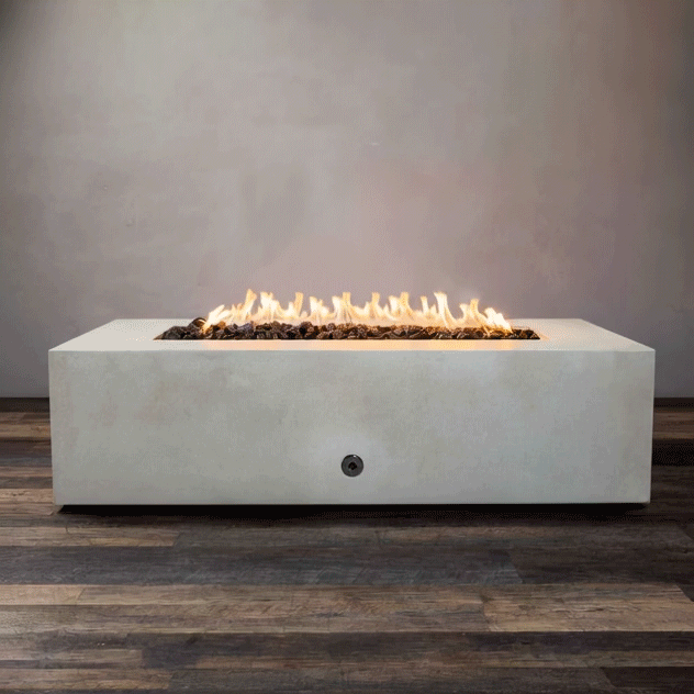 A studio presentation displays a rectangular fire pit in a crisp white finish, resting on a dark wood floor. Bright yellow flames burn continuously along the linear fire pit burner, contrasting with the smooth texture of the concrete fire pit. The visible chrome key valve on the side panel indicates the manual control of this natural gas fire pit unit, demonstrating its clean aesthetic and functionality.