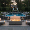 A grand poolside terrace features a white luxury fire pit surrounded by elegant sofas and armchairs. The setting is framed by a manicured lawn, pool house, and mature trees at sunset, creating a sophisticated backyard oasis. The warm glow of the outdoor fire pit anchors the family gathering fire pit area, seamlessly integrating with the estate's formal landscape design.