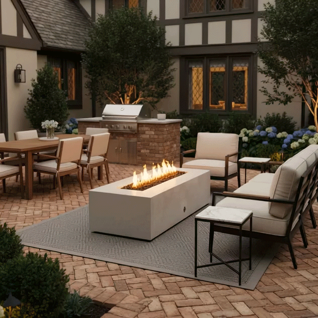 A charming brick courtyard features a white patio fire pit surrounded by cushioned seating and an outdoor dining area. The setting includes an outdoor kitchen and fireplace, creating a complete outdoor entertaining space. The steady flames of the propane fire pit provide warmth and ambiance, enhancing the cozy backyard fire pit atmosphere in a traditional garden setting.