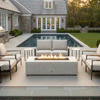 A classic shingle-style home features a white fire table on a bluestone patio next to a swimming pool. Surrounded by white armchairs and sofas, the fire feature serves as a focal point for the backyard. The clean-burning flames illustrate the benefits of a smokeless fire pit, with the gas fire pit adding warmth and elegance to the coastal estate setting.