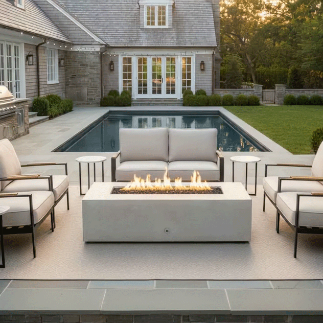 A classic shingle-style home features a white fire table on a bluestone patio next to a swimming pool. Surrounded by white armchairs and sofas, the fire feature serves as a focal point for the backyard. The clean-burning flames illustrate the benefits of a smokeless fire pit, with the gas fire pit adding warmth and elegance to the coastal estate setting.