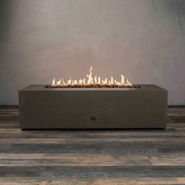 A long, low brown concrete fire pit with crisp rectangular sides sits centered on wood-look flooring in an otherwise empty room, its recessed channel filled with uneven dark lava rocks while a continuous ribbon of golden flames flickers along the linear burner, highlighting the strong horizontal shape of this concrete fire pit and the slim proportions of a modern rectangular fire pit set against a plain gray wall.