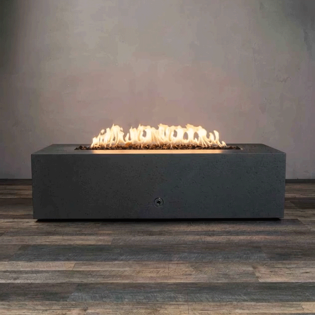 A dark graphite concrete fire pit with a long, low rectangular shape sits on wood-look decking in a bare room, its narrow trough filled with rough lava rocks while a continuous line of yellow flames flickers along the center, emphasizing this concrete fire pit as a sleek rectangular fire pit and linear fire pit that adds a strong horizontal glow against the plain gray backdrop.