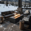 A graphite concrete fire pit burns on a covered patio overlooking a snowy ski slope, its rectangular opening filled with rocks and bright flames as skiers move in the distance, while gray cushioned sofas, armchairs, fur throws, stump tables, and lanterns circle the hearth, creating an outdoor fire pit that functions as a rectangular fire pit and cozy backyard fire pit for warming up after a day on the mountain.