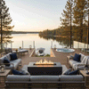 On a multitiered lakeside deck enclosed by glass railings, a graphite concrete fire pit glows at the center of gray cushioned sectionals and loungers, its linear burner casting warm light across the boards while lanterns, side tables, and a round hot tub frame views of still water and tall pines at sunset, showcasing an outdoor fire pit used as a sleek rectangular fire pit and modern fire pit within a tranquil backyard oasis.