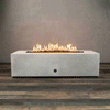 A long white concrete fire pit with crisp rectangular edges sits alone on a wood-look floor, its recessed linear burner filled with dark lava rocks as a continuous row of yellow flames rises and sways against a plain gray wall, emphasizing the low, boxy shape of this concrete fire pit and the strong horizontal line of the rectangular fire pit that stretches nearly the full length of the frame.