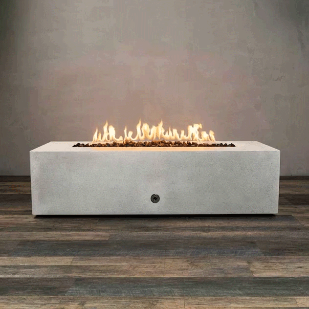 A long white concrete fire pit with crisp rectangular edges sits alone on a wood-look floor, its recessed linear burner filled with dark lava rocks as a continuous row of yellow flames rises and sways against a plain gray wall, emphasizing the low, boxy shape of this concrete fire pit and the strong horizontal line of the rectangular fire pit that stretches nearly the full length of the frame.