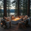 On a raised deck nestled among tall pines above a calm lake, a white rectangular concrete fire pit burns with a bright linear flame at the center of wooden lounge chairs and a sofa layered with blankets and pillows, lanterns glowing at the edges and a hammock hanging between trees behind, creating an outdoor fire pit that feels like a cozy backyard fire pit and natural family gathering fire pit in the forest at dusk.