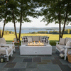 A white concrete fire pit with a long rectangular opening burns in the middle of a slate patio bordered by leafy trees, its flames rising above dark stones while matching white outdoor sofas and armchairs with plaid pillows form a U-shaped seating area, overlooking a wide green field and distant lake, presenting a patio fire pit that anchors relaxed outdoor entertaining and turns the lakeside setting into a casual backyard oasis.