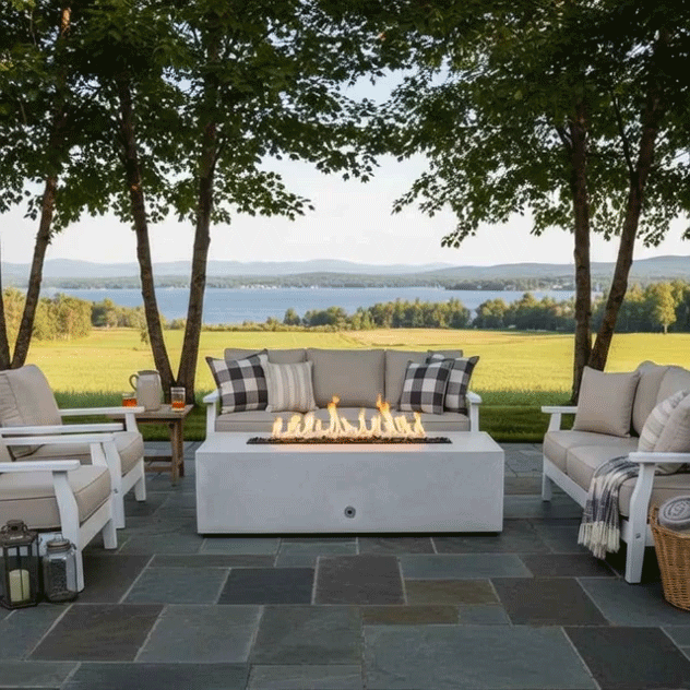 A white concrete fire pit with a long rectangular opening burns in the middle of a slate patio bordered by leafy trees, its flames rising above dark stones while matching white outdoor sofas and armchairs with plaid pillows form a U-shaped seating area, overlooking a wide green field and distant lake, presenting a patio fire pit that anchors relaxed outdoor entertaining and turns the lakeside setting into a casual backyard oasis.
