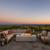 On a broad flagstone terrace surrounded by wildflower beds and open fields, a white rectangular concrete fire pit glows with a steady linear flame at the center of an L-shaped sectional and armchairs, lanterns resting on nearby tables and a telescope aimed toward the fading sunset sky, depicting an outdoor fire pit styled as a modern fire pit and spacious family gathering fire pit for evenings spent under the changing colors of the horizon.