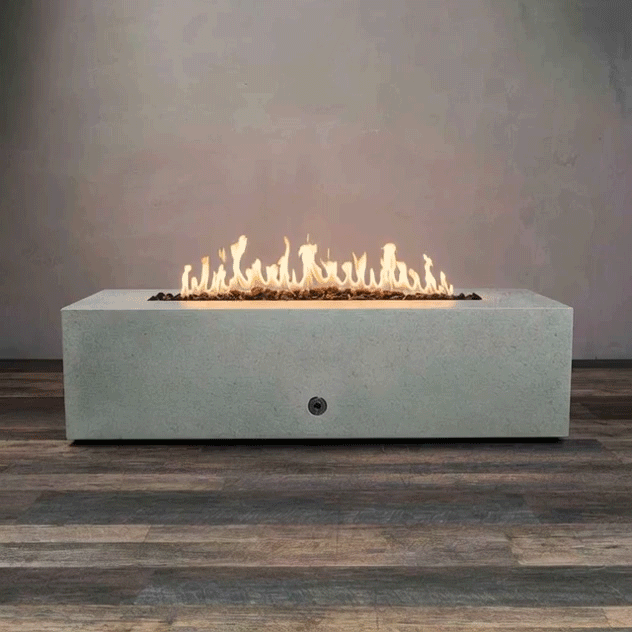 A long, low concrete fire pit in a pale gray finish sits centered on a wood-look floor against a plain wall, its recessed rectangular opening filled with uneven dark lava rocks while a continuous ribbon of yellow flames ripples along the burner, drawing the eye to the simple boxy shape of this concrete fire pit and the strong horizontal line of a minimalist rectangular fire pit with a linear fire pit flame pattern.