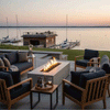 On a stone terrace overlooking a calm lake and small marina at sunset, a pale concrete fire pit burns between four dark-cushioned wooden lounge chairs, its long rectangular trough packed with lava rocks and a bright line of flames, turning this concrete fire pit into an inviting outdoor fire pit and rectangular fire pit focal point for relaxed outdoor entertaining with drinks set on small side tables around the seating circle.