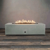 A long, low concrete fire pit in a pale gray finish stretches across the center of the frame, its rectangular top cut with a narrow trough filled with dark lava rocks while an even line of yellow flames flickers and dances along the length of the burner, the simple boxy base with a small round control resting on wood-look flooring against a plain wall and emphasizing this concrete fire pit as a minimalist rectangular and linear fire pit in a bare studio setting.