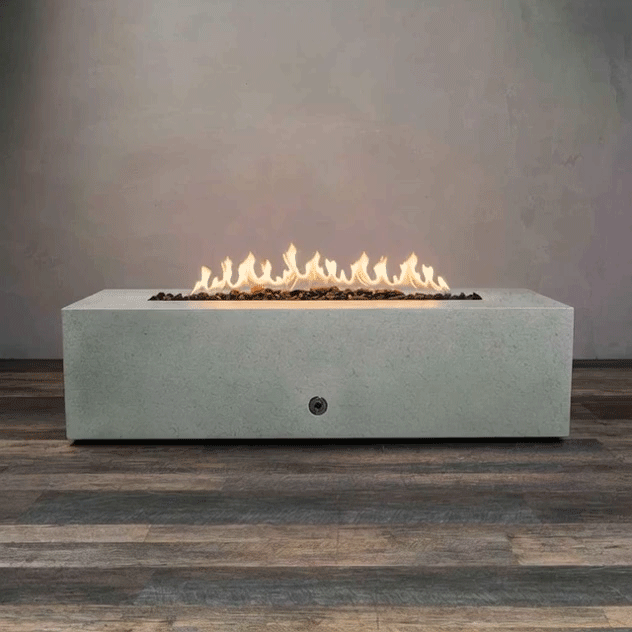 A long, low concrete fire pit in a pale gray finish stretches across the center of the frame, its rectangular top cut with a narrow trough filled with dark lava rocks while an even line of yellow flames flickers and dances along the length of the burner, the simple boxy base with a small round control resting on wood-look flooring against a plain wall and emphasizing this concrete fire pit as a minimalist rectangular and linear fire pit in a bare studio setting.