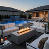 On a modern backyard deck at dusk, a sleek concrete fire pit sits between white cushioned sofas beside a glowing pool and an illuminated outdoor kitchen, its long rectangular opening filled with dark stones as a row of flames rises and reflects on the surrounding metal lanterns and drink glasses, turning the piece into a low fire table that anchors relaxed outdoor entertaining in a carefully designed lounge-style backyard space.