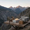 A wooden platform deck juts out over a steep rocky canyon, enclosed by slim cable railing and furnished with a cushioned sofa, two armchairs, and a chunky wood side table, all focused on a pale concrete fire pit whose linear burner sends up a steady row of flames, presenting an outdoor fire pit with a rectangular profile and modern fire pit styling that contrasts the smooth surfaces with the rugged mountain landscape beyond.