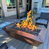 Starfire Designs 60" x 32" Copper Canyon gas fire pit with a glass wind guard, displayed on a modern patio with vibrant flames and a hammered copper finish