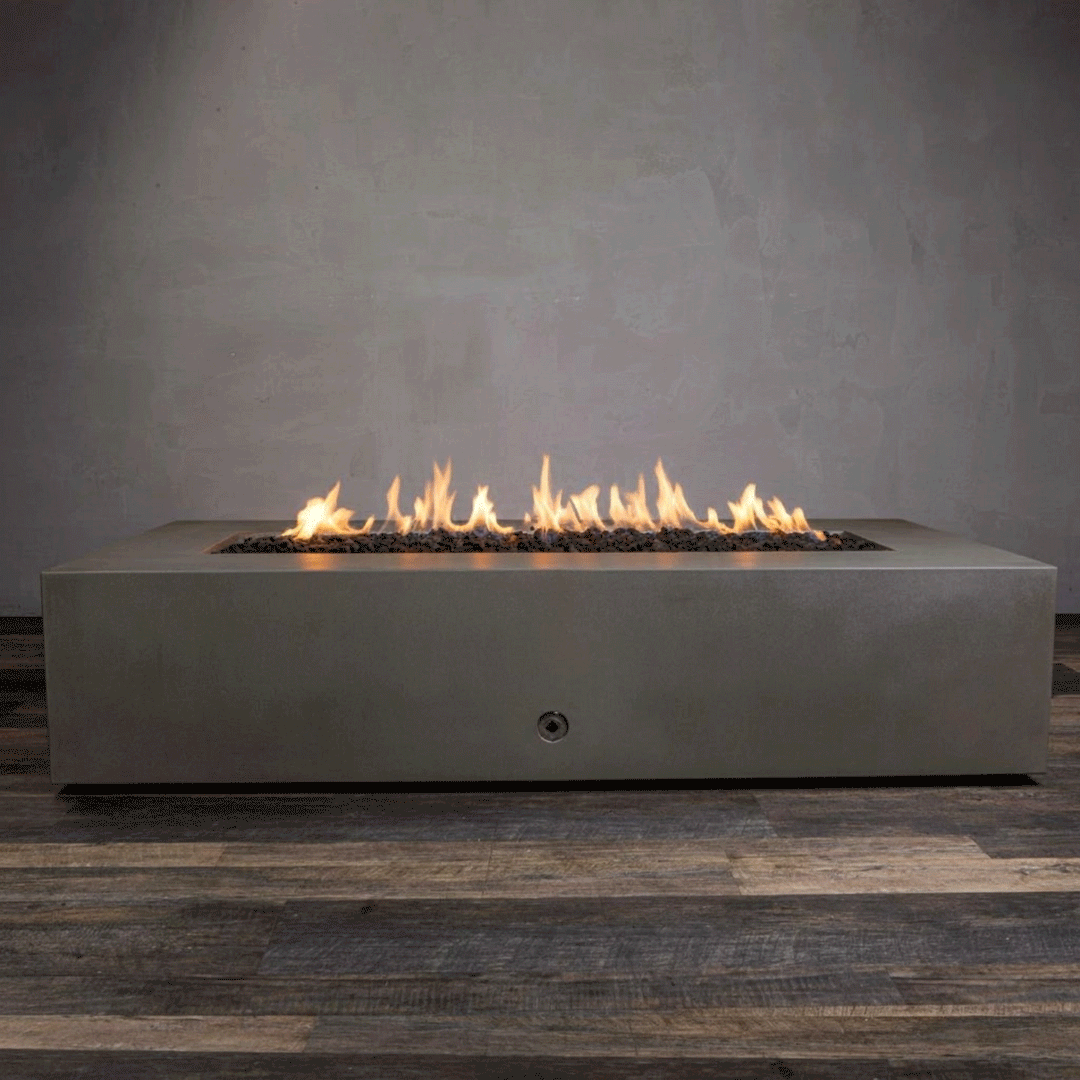 A studio presentation displays a large rectangular fire pit in a natural brown finish, resting on a dark wood floor. Bright yellow flames burn continuously along the linear fire pit burner, rising from a bed of black lava rocks. The visible chrome key valve on the side indicates the manual control of this concrete fire pit, highlighting the earth-tone texture and clean operation of a natural gas fire pit connection.