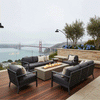 A brown modern fire pit sits on a hillside deck overlooking the Golden Gate Bridge and ocean on a misty day. Surrounded by dark grey sofas and hydrangeas, the outdoor fire pit anchors the seating area. The bright flames of the gas fire pit add warmth and contrast to the cool, foggy backdrop, serving as a striking fire feature in a scenic location.
