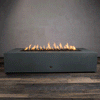 A studio presentation highlights a large rectangular fire pit in a matte graphite finish, resting on a dark wood floor. Bright orange flames flicker continuously along the linear fire pit burner, rising from a bed of black lava rocks. The visible chrome key valve on the side panel indicates the manual control of this concrete fire pit, showcasing the sleek profile and steady combustion of a natural gas fire pit connection.