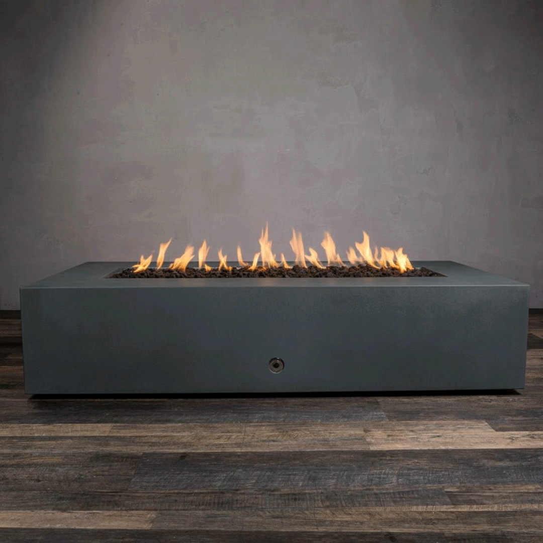 A studio presentation highlights a large rectangular fire pit in a matte graphite finish, resting on a dark wood floor. Bright orange flames flicker continuously along the linear fire pit burner, rising from a bed of black lava rocks. The visible chrome key valve on the side panel indicates the manual control of this concrete fire pit, showcasing the sleek profile and steady combustion of a natural gas fire pit connection.