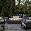 A charming courtyard patio features a graphite outdoor fire pit surrounded by an elegant sofa and armchairs. Ivy-covered brick walls and mature trees frame the scene, creating a lush backyard oasis. The warm glow of the fire table illuminates the stone pavers, serving as an inviting family gathering fire pit area for evening relaxation.