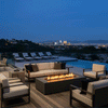A graphite luxury fire pit anchors a spacious wooden deck overlooking a city skyline at night. Positioned near an infinity pool and lounge chairs, the modern fire pit is surrounded by a large sofa and armchairs. The steady amber flames of the gas fire pit create a sophisticated atmosphere for outdoor entertaining against a panoramic urban backdrop.