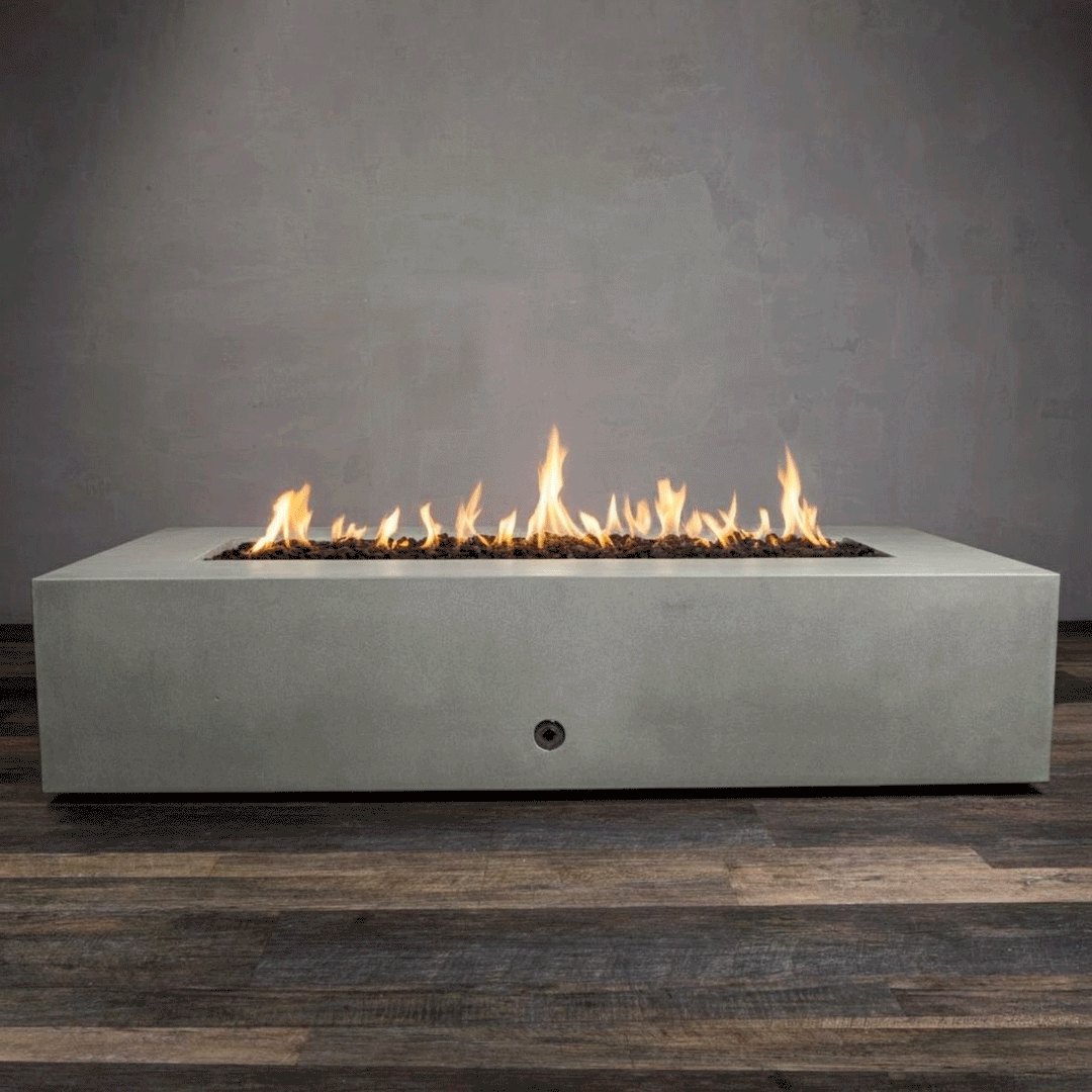 A studio presentation highlights a large rectangular fire pit in a natural gray finish, resting on a dark wood floor. Bright yellow flames burn continuously along the linear fire pit burner, rising from a bed of black lava rocks. The visible chrome key valve on the side panel indicates the manual control of this concrete fire pit, showcasing the clean lines and steady operation of a natural gas fire pit connection.