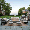 A spacious flagstone patio features a gray luxury fire pit surrounded by cushioned armchairs and sofas. The setting overlooks a manicured lawn and ocean horizon at twilight, creating an elegant outdoor entertaining area. The warm glow of the propane fire pit anchors the seating arrangement, serving as a sophisticated patio fire pit centerpiece for the coastal estate.