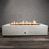 A studio presentation highlights a large rectangular fire pit in a crisp white finish, resting on a dark wood floor. Bright orange flames burn steadily along the linear fire pit burner, contrasting with the smooth texture of the concrete fire pit. The visible chrome key valve on the side panel indicates the manual control of this natural gas fire pit unit, demonstrating its clean design and functionality.