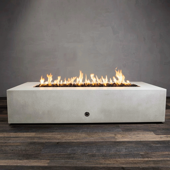 A studio presentation highlights a large rectangular fire pit in a crisp white finish, resting on a dark wood floor. Bright orange flames burn steadily along the linear fire pit burner, contrasting with the smooth texture of the concrete fire pit. The visible chrome key valve on the side panel indicates the manual control of this natural gas fire pit unit, demonstrating its clean design and functionality.