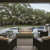 A covered patio features a white gas fire pit positioned between a sofa and armchairs. The setting overlooks a backyard swimming pool and mature trees, creating a private backyard oasis. The warm glow of the fire provides ambiance for a family gathering fire pit area, seamlessly integrating with the outdoor living space.