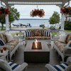On a covered porch with ornate trim overlooking a quiet waterfront street and distant lake, a taupe concrete fire pit burns gently between white wicker loveseats and chairs upholstered in floral cushions with striped pillows, hanging baskets and lanterns scattered around, depicting a patio fire pit that acts as a cozy backyard fire pit and intimate family gathering fire pit where people can linger with iced tea while horse-drawn carriages pass outside.