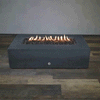 A low graphite concrete fire pit with a long rectangular shape sits centered on wood-look flooring against a plain gray wall, its recessed trough packed with dark, uneven lava rocks while a steady row of yellow flames flickers along the burner, emphasizing the minimalist silhouette of this concrete fire pit and the elongated profile of a sleek rectangular fire pit with a linear fire pit flame pattern running almost the full length of the top surface.