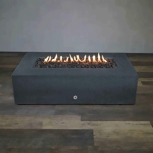 A low graphite concrete fire pit with a long rectangular shape sits centered on wood-look flooring against a plain gray wall, its recessed trough packed with dark, uneven lava rocks while a steady row of yellow flames flickers along the burner, emphasizing the minimalist silhouette of this concrete fire pit and the elongated profile of a sleek rectangular fire pit with a linear fire pit flame pattern running almost the full length of the top surface.