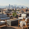 A graphite concrete fire pit with a long rectangular opening glows at the center of a rooftop deck, its flames rising above lava rocks while pale gray cushioned chairs and sofas with wooden arms form a ring around it, potted grasses behind and a city skyline with distant mountains beyond the railing, presenting an outdoor fire pit that functions as a modern rectangular fire pit and concrete fire pit focal point for casual outdoor entertaining above the streets.