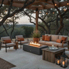 Under a wooden pergola wrapped with string lights and framed by twisting trees, a graphite concrete fire pit burns with a long ribbon of flame between cushioned chairs and a sofa accented with rust-colored pillows, a rustic wood coffee table and lanterns nearby, illustrating a patio fire pit that doubles as a rectangular fire pit and concrete fire pit centerpiece for cozy backyard fire pit gatherings in a relaxed, tree-lined outdoor living space.