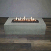 A long, pale gray concrete fire pit with clean rectangular lines sits centered on wood-look flooring in a bare studio, its recessed channel filled with uneven dark lava rocks while a steady row of yellow flames ripples along the burner, drawing attention to the low, boxy form of this concrete fire pit and the elongated shape of a minimalist rectangular fire pit with a linear fire pit flame pattern.