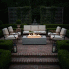 In a formal brick courtyard framed by clipped boxwood hedges and lattice screens, a light gray concrete fire pit stretches between cushioned metal armchairs and a sofa, its linear flames rising above dark stones while lanterns and a tiered fountain glow softly in the background, turning the space into an elegant patio fire pit setting that feels like a refined concrete fire pit and intimate fire feature for relaxed outdoor entertaining.