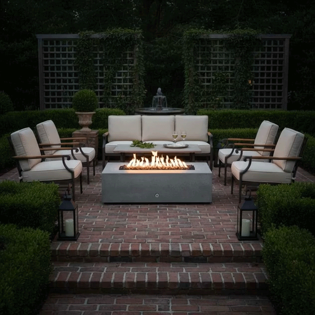 In a formal brick courtyard framed by clipped boxwood hedges and lattice screens, a light gray concrete fire pit stretches between cushioned metal armchairs and a sofa, its linear flames rising above dark stones while lanterns and a tiered fountain glow softly in the background, turning the space into an elegant patio fire pit setting that feels like a refined concrete fire pit and intimate fire feature for relaxed outdoor entertaining.