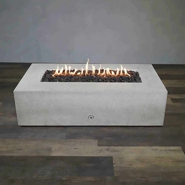 A long white concrete fire pit with crisp rectangular sides sits centered on a wood-look floor against a plain gray wall, its recessed trough filled with dark lava rocks while a steady row of yellow flames ripples along the linear burner, emphasizing the low minimalist form of this concrete fire pit and the elongated shape of a modern rectangular fire pit in an otherwise empty studio setting.