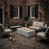 In a brick-walled courtyard between traditional windows with dark shutters, a white rectangular concrete fire pit glows on weathered herringbone pavers, its line of flames rising above lava rocks while cushioned metal chairs and a small sofa surround it, string lights and lanterns adding extra sparkle, creating a patio fire pit that turns the enclosed space into a cozy backyard fire pit for relaxed outdoor entertaining.