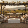Under a wooden pergola draped with late-season vines, a white concrete fire pit burns at the center of gravel seating lined with soft neutral chairs and sofas, its rectangular opening filled with stones and a bright linear flame while a long farm table and rolling vineyard hills stretch out behind, forming an outdoor fire pit that serves as a low rectangular fire pit focal point in a relaxed, vineyard-style backyard oasis.