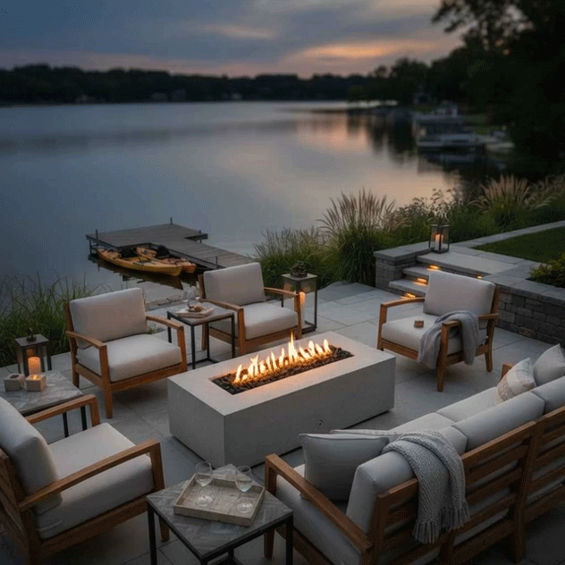 On a stepped stone patio beside a calm lake at dusk, a white rectangular concrete fire pit glows between matching wood-framed lounge chairs and a sofa, its continuous row of flames reflecting softly on the water while lanterns, grasses, and a small dock with kayaks frame the scene, showing an outdoor fire pit used as a cozy backyard fire pit and inviting family gathering fire pit as the sky darkens.