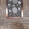 Close-up of a rectangular oil-rubbed bronze fire pit burner filled with natural lava rocks and smooth stones, set against a rustic wood tabletop for a contemporary outdoor fire feature.