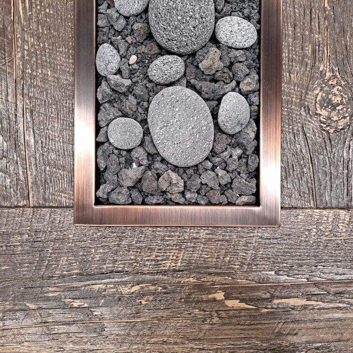 Close-up of a rectangular oil-rubbed bronze fire pit burner filled with natural lava rocks and smooth stones, set against a rustic wood tabletop for a contemporary outdoor fire feature.