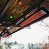 Close-up of two Bromic Heating Tungsten Smart-Heat Electric Heaters in black, mounted under a rustic patio roof with string lights, delivering radiant heat for outdoor comfort.